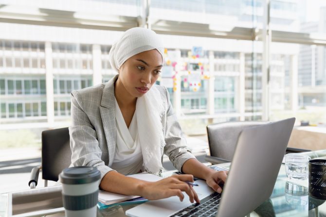 Front view of businesswoman in hijab working on laptop at desk in a modern office. Modern corporate start up new business concept with entrepreneur working hard